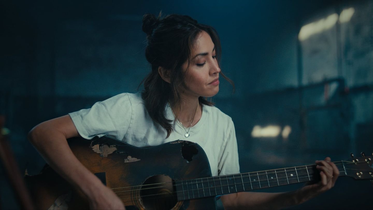 A woman plays a worn acoustic guitar in a blue lit warehouse